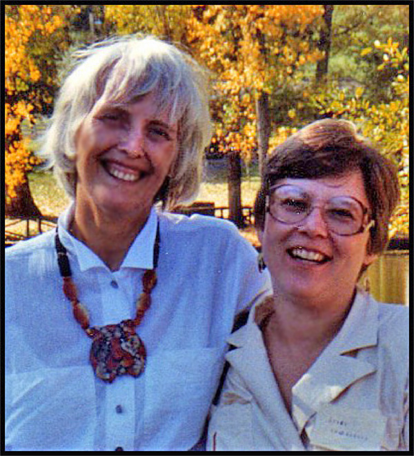 Dona and her mother are smiling and holding each other, standing in front of a pond with autumn-colored trees behind the pond.